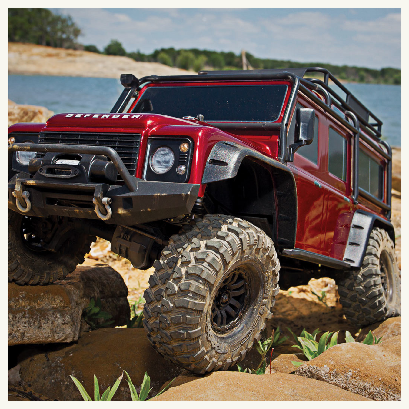 Red Land Rover Defender crawling on rocky shore with lake in the background